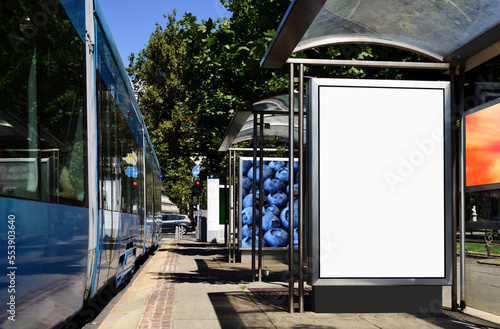 blank white lightbox ad panel on the side. poster and advertising billboard sign. mockup base. business communication placeholder. blue and red tram in closeup view. streetcar stop. soft background