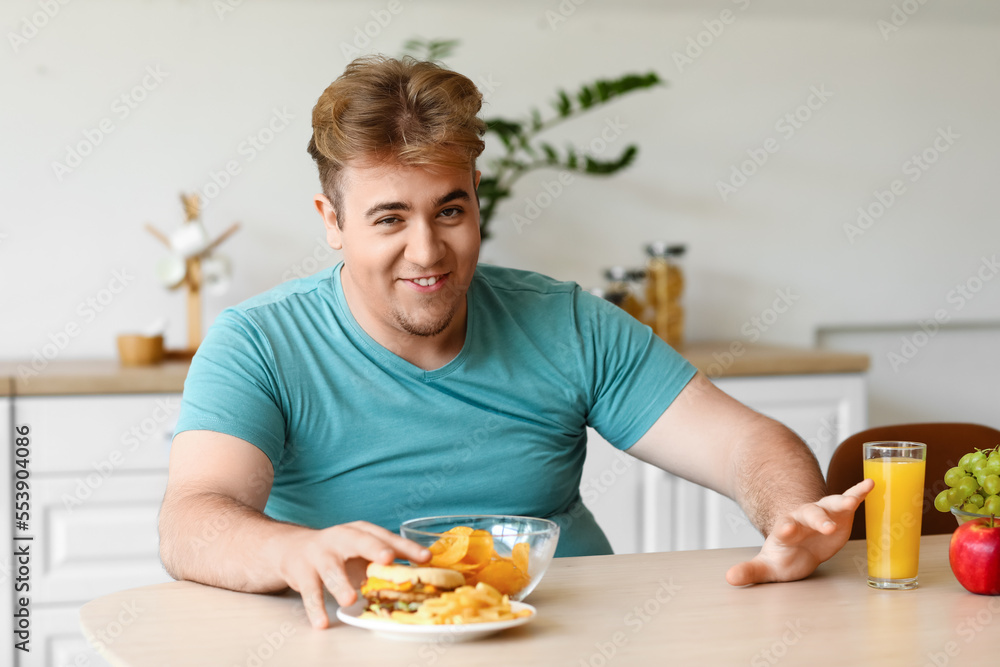 Young overweight man rejecting healthy food at table in kitchen Young overweight man rejecting healthy food at table in kitchen
