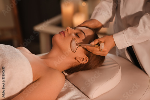 Pretty young woman receiving face massage with roller in beauty salon