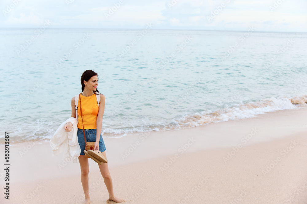 Woman smile with teeth after swimming in the ocean with a backpack in a wet yellow tank top and denim shorts walks along the beach, summer vacation on an island by the ocean in Bali sunset