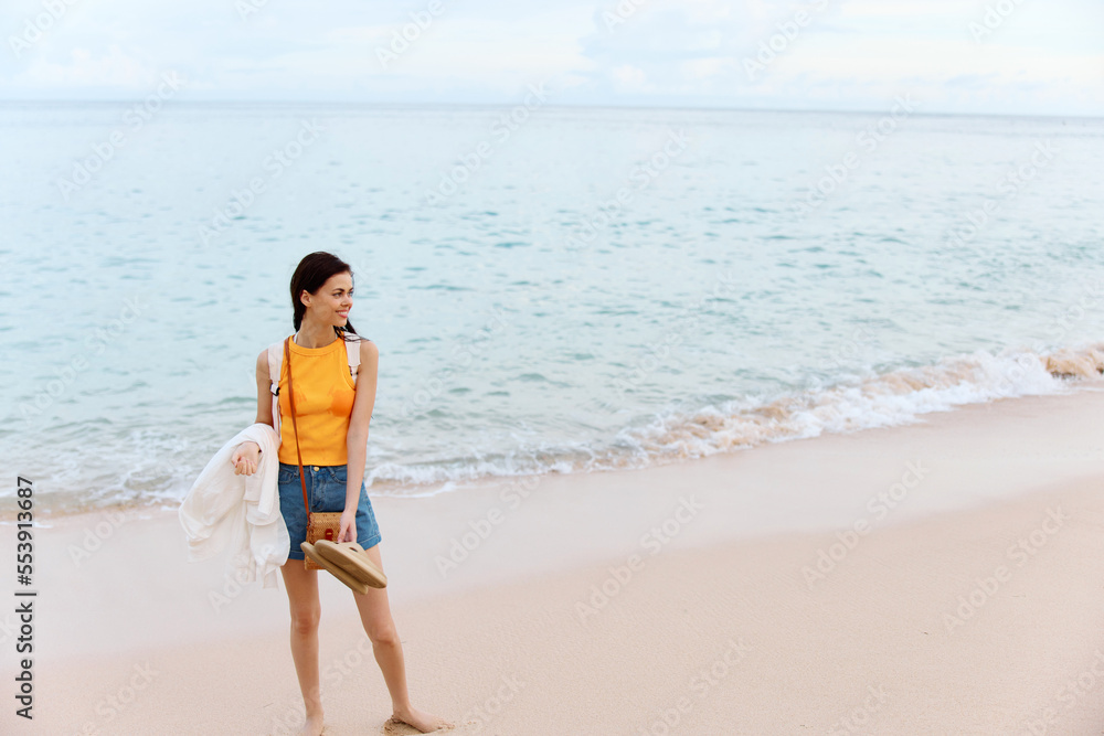 Woman smile with teeth after swimming in the ocean with a backpack in a wet yellow tank top and denim shorts walks along the beach, summer vacation on an island by the ocean in Bali sunset Woman smile with teeth after swimming in the ocean with a backpack in a wet yellow tank top and denim shorts walks along the beach, summer vacation on an island by the ocean in Bali sunset