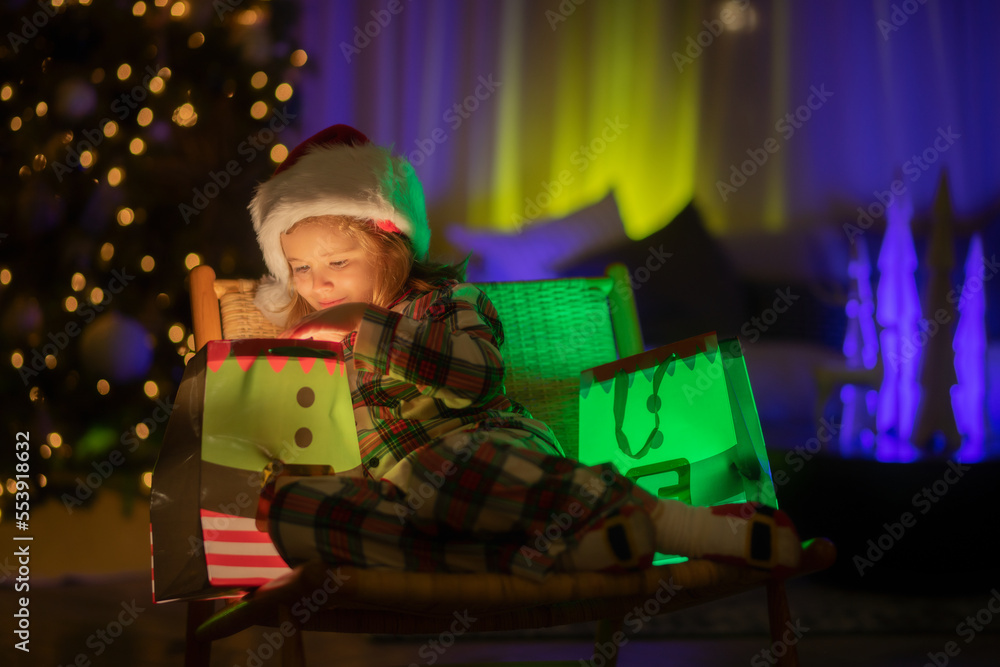Kid with present gift with magic light. Lighting present gift bag. Child in santa hat on front of night Christmas tree home background on Christmas Eve.