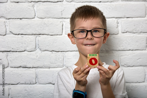 Handsome boy in a white T-shirt stands against a brick wall with a wooden cube in his hands. Little boy with a smart watch on his arm holds a wooden toy in the form of a cube to learn the alphabet