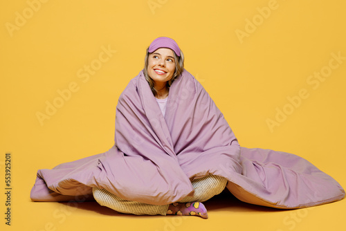 Full body young happy fun woman she wears purple pyjamas jam sleep eye mask rest relax at home sit wrap covered blanket duvet look aside on area isolated on plain yellow background. Night nap concept.