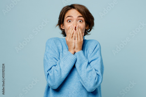 Young astonished amazed happy winner caucasian woman in knitted sweater look camera cover mouth with hand isolated on plain pastel light blue cyan background studio portrait People lifestyle concept
