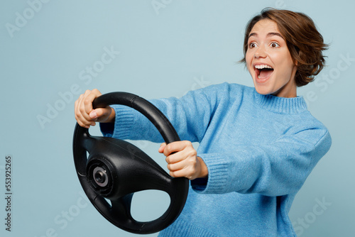 Side view young excited cheerful fun woman in knitted sweater look camera hold steering wheel driving car isolated on plain pastel light blue cyan background studio portrait People lifestyle concept