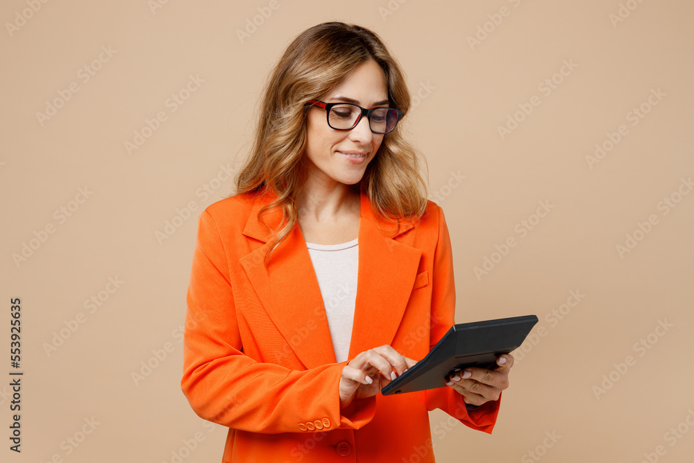 Young smiling fun successful employee business woman corporate lawyer 30s in classic formal orange suit glasses work in office hold in hand using calculator isolated on plain beige background studio