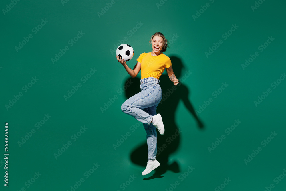 Side view full body young woman fan in basic yellow t-shirt cheer up support football sport team hold in hand soccer ball watch tv live stream jump high clench fist isolated on dark green background Side view full body young woman fan in basic yellow t-shirt cheer up support football sport team hold in hand soccer ball watch tv live stream jump high clench fist isolated on dark green background