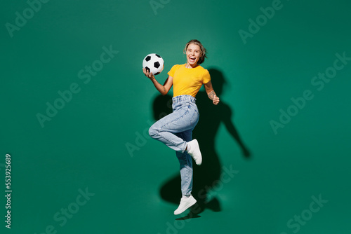 Fototapeta Naklejka Na Ścianę i Meble -  Side view full body young woman fan in basic yellow t-shirt cheer up support football sport team hold in hand soccer ball watch tv live stream jump high clench fist isolated on dark green background