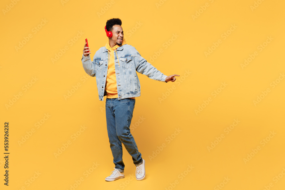 Full body side view fun young man of African American ethnicity wear denim jacket hoody headphones listen to music use mobile cell phone isolated on plain yellow background People lifestyle concept Full body side view fun young man of African American ethnicity wear denim jacket hoody headphones listen to music use mobile cell phone isolated on plain yellow background People lifestyle concept