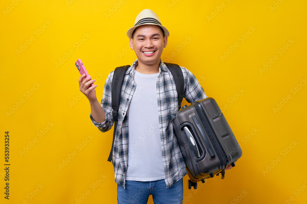 Cheerful young traveler tourist Asian man in casual clothes hat with backpack carrying suitcase in hand and mobile phone isolated on yellow background. Air flight journey concept
