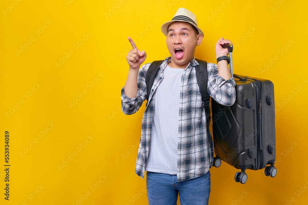 Excited young traveler tourist Asian man 20 years old wears casual clothes hat with backpack carrying suitcase, pointing finger and looking aside on yellow background. Air flight journey concept