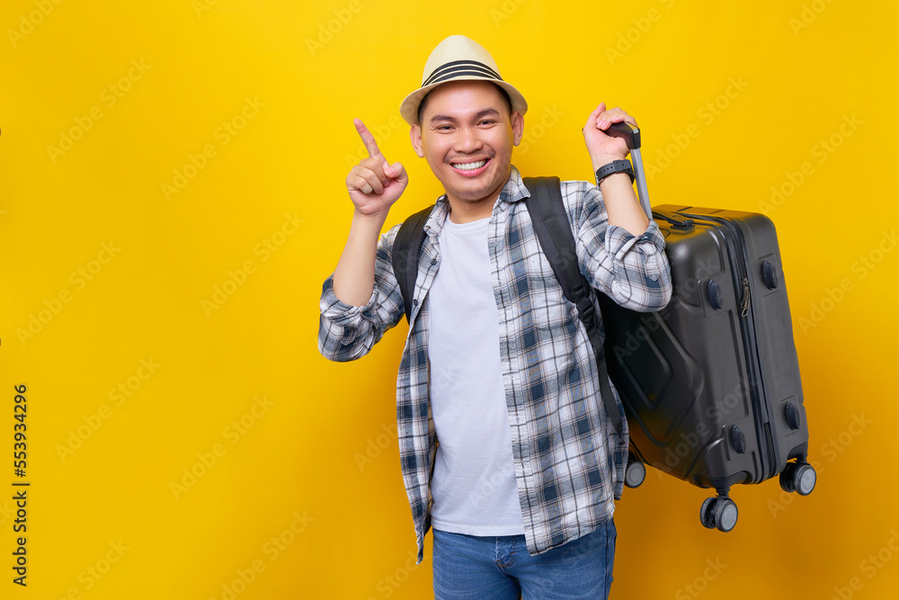Smiling young traveler tourist Asian man wearing casual clothes hat with backpack carrying suitcase, pointing the finger at copy space isolated on yellow background. Air flight journey concept