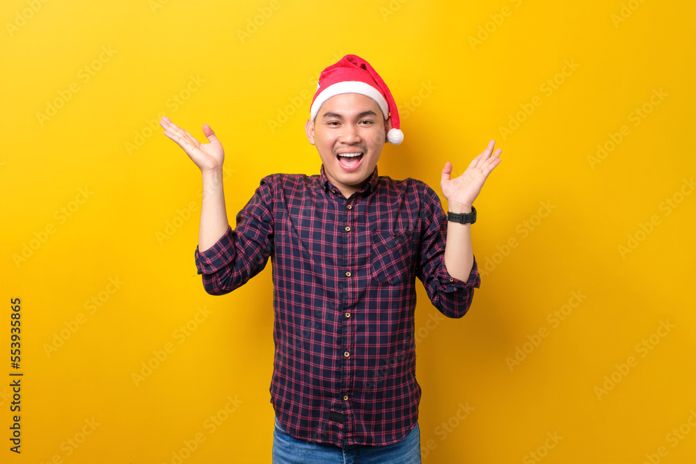 Surprised young Asian man in Santa hat raising hands and looking at camera with excitement expression on yellow studio background. Happy New Year 2023 celebration merry holiday concept Surprised young Asian man in Santa hat raising hands and looking at camera with excitement expression on yellow studio background. Happy New Year 2023 celebration merry holiday concept