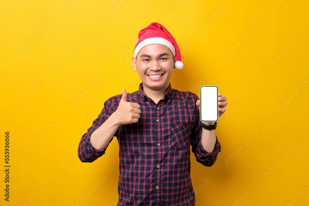 Cheerful young Asian man in Santa hat holding mobile phone with blank screen, showing thumb up gesture over yellow studio background. Happy New Year 2023 celebration merry holiday concept Cheerful young Asian man in Santa hat holding mobile phone with blank screen, showing thumb up gesture over yellow studio background. Happy New Year 2023 celebration merry holiday concept