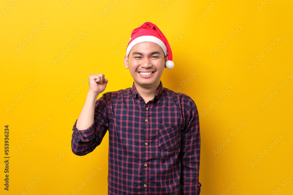 Excited young Asian man in Santa hat raising hands up, celebrating success on yellow studio background. Happy New Year 2023 celebration merry holiday concept