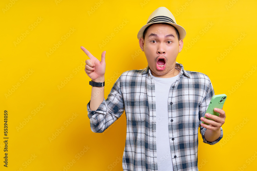 Amazed traveler tourist Asian man in casual clothes hat holding mobile phone and pointing at copy space on yellow background. Passenger traveling on weekends. Air flight journey concept Amazed traveler tourist Asian man in casual clothes hat holding mobile phone and pointing at copy space on yellow background. Passenger traveling on weekends. Air flight journey concept