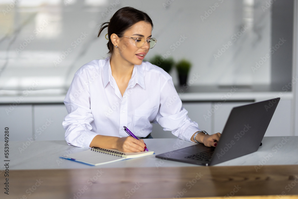 Young woman sit at table with laptop in kitchen holds pen, take notes on paper, writes letter. Young woman sit at table with laptop in kitchen holds pen, take notes on paper, writes letter.