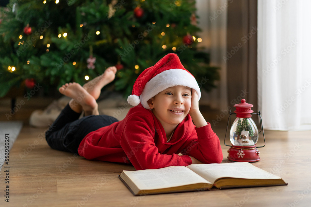 A little boy in a Santa Claus hat lies on the floor and reads a book with fairy tales