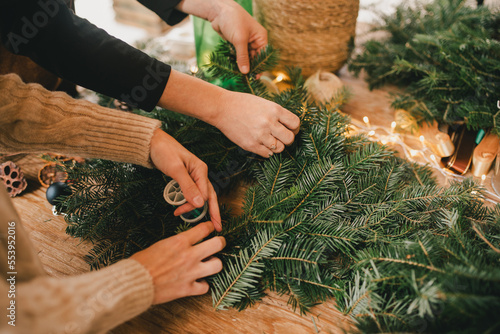 Two women making natural Christmas wreath using pine branches and festive holiday decorations.