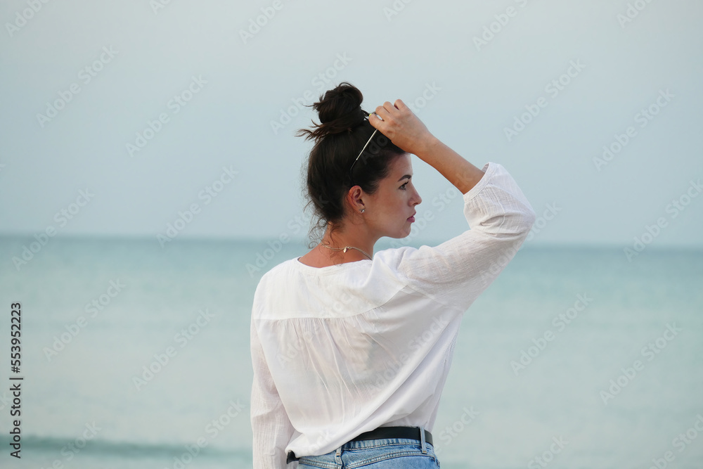Slim girl walking by the sea. Summer lifestyle image of happy woman walking on the beach. Pretty young woman with long hair in a white shirt and denim shorts. Slim girl walking by the sea. Summer lifestyle image of happy woman walking on the beach. Pretty young woman with long hair in a white shirt and denim shorts.