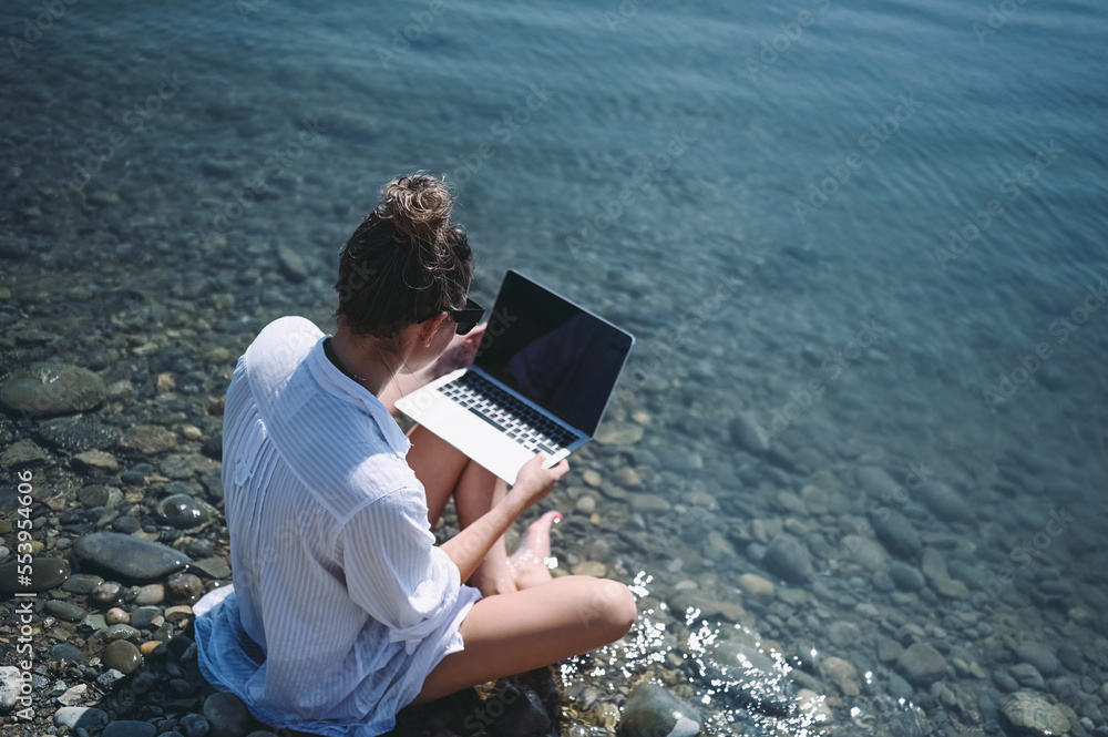 Young woman freelancer traveler wearing white shirt anywhere working online outdoors using laptop enjoying lake view. Happy female downshifter in sunglasses holding computer at sea coastline 