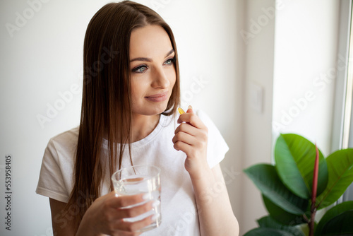 Woman taking vitamin pill with glass of fresh water indoors.