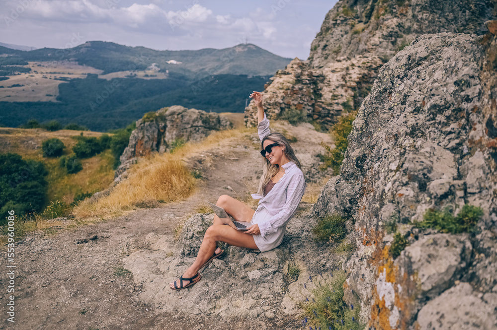 Young woman freelancer traveler working online anywhere outdoors using laptop enjoying mountain peak view. Happy female downshifter in sunglasses holding computer in unusual workplace at summer