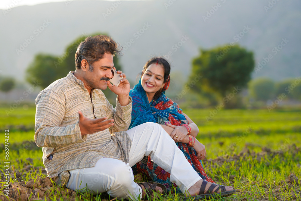 Indian farmer sitting with wife and talking on smartphone at agriculture field. Indian farmer sitting with wife and talking on smartphone at agriculture field.