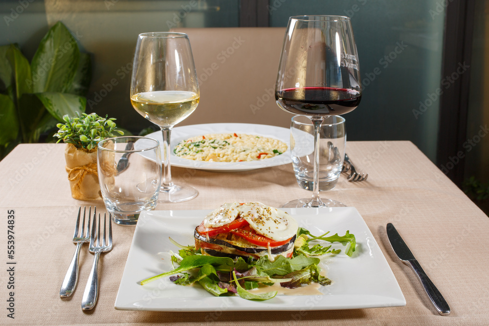 Tavolo di un ristorante elegante apparecchiato con gusto e imbandito con un antipasto, un primo piatto, un calice di vino bianco e un calice di vino rosso