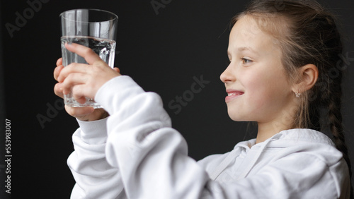Girl drinks clean water. Positive child is holding transpired glass cup with freshwater and smiling indoors. Healthy food child concept of children's happiness.