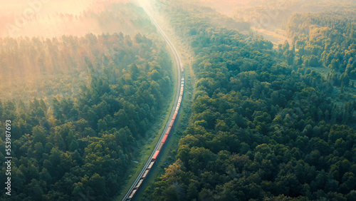 Cargo Train in summer morning forest at fog sunrise. Aerial view of moving freight train in forest. Morning mist landscape with train, railroad, foggy trees. Top aerial drone view near railway. © Andrii Chagovets