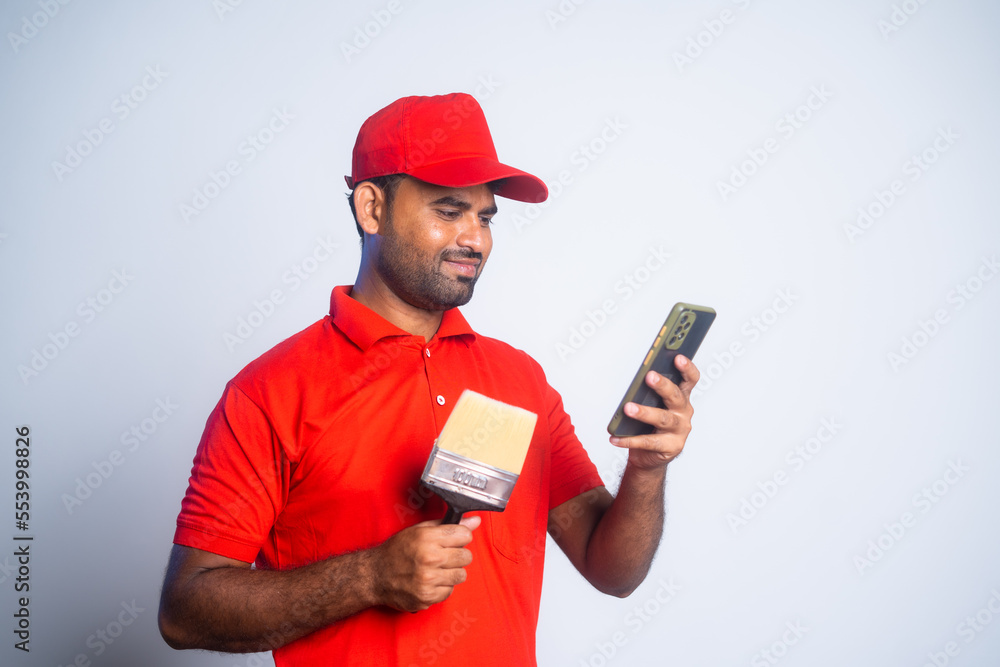 Happy painter busy using mobile phone by holding paint brush at construction site - concept of social media, technology and relaxation Happy painter busy using mobile phone by holding paint brush at construction site - concept of social media, technology and relaxation