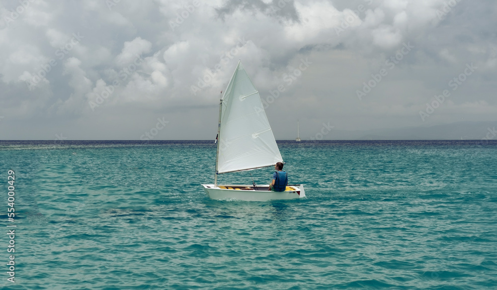 Lonely sailor on training sailing pram optimist education boat in the sea in Greece, water background and cloudy sky - obrazy, fototapety, plakaty 