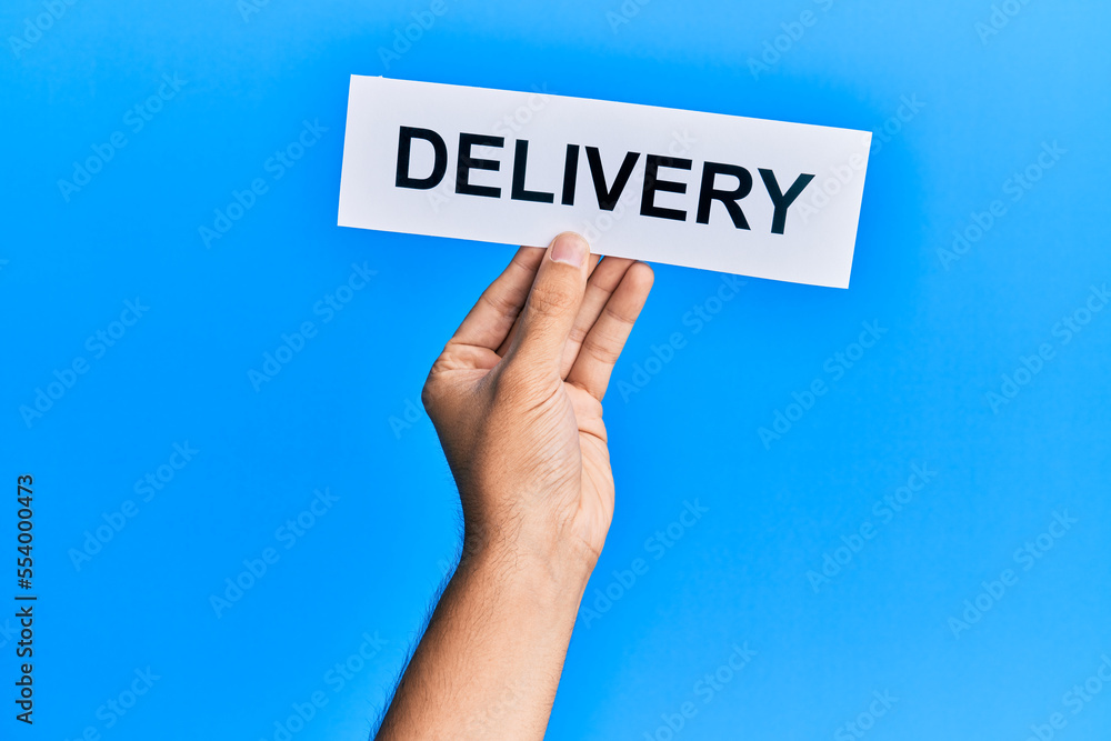 Hand of caucasian man holding paper with delivery word over isolated blue background Hand of caucasian man holding paper with delivery word over isolated blue background