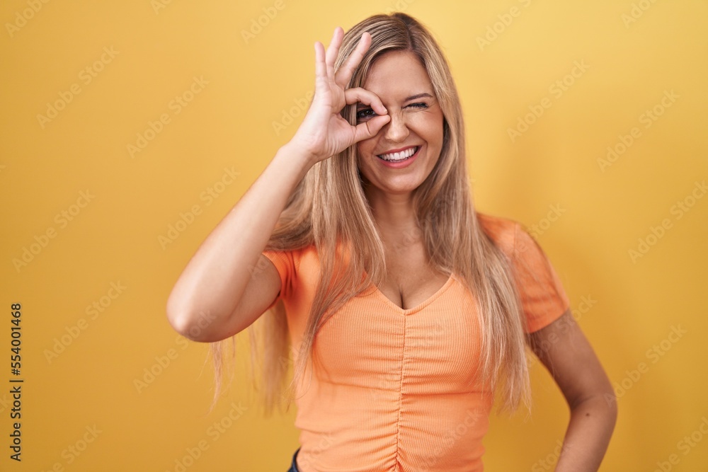 Young woman standing over yellow background doing ok gesture with hand smiling, eye looking through fingers with happy face. Young woman standing over yellow background doing ok gesture with hand smiling, eye looking through fingers with happy face.