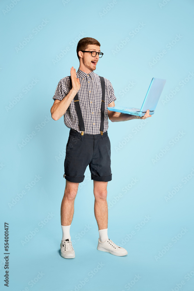 Portrait of young man in checkered shirt, shorts and suspenders having video call on laptop isolated over blue background. Internet meeting