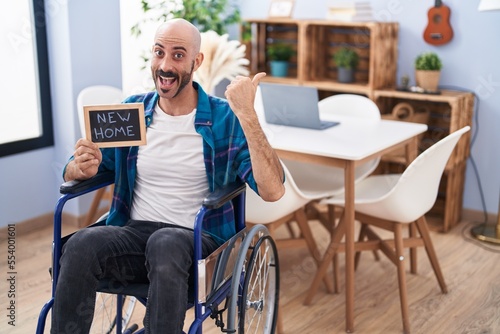 Hispanic man with beard sitting on wheelchair at new home pointing thumb up to the side smiling happy with open mouth