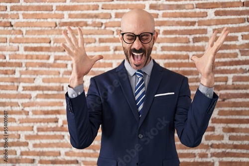 Bald man with beard wearing business clothes and glasses celebrating mad and crazy for success with arms raised and closed eyes screaming excited. winner concept