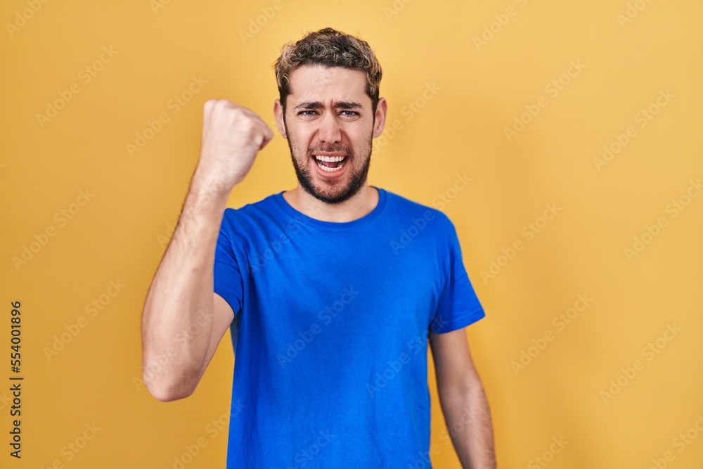 Hispanic man with beard standing over yellow background angry and mad raising fist frustrated and furious while shouting with anger. rage and aggressive concept. Hispanic man with beard standing over yellow background angry and mad raising fist frustrated and furious while shouting with anger. rage and aggressive concept.