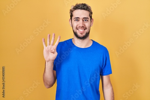 Hispanic man with beard standing over yellow background showing and pointing up with fingers number four while smiling confident and happy.