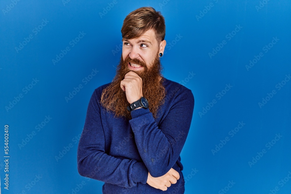 Redhead man with long beard wearing casual blue sweater over blue background thinking worried about a question, concerned and nervous with hand on chin Redhead man with long beard wearing casual blue sweater over blue background thinking worried about a question, concerned and nervous with hand on chin