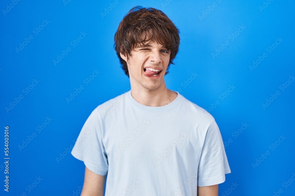 Hispanic young man standing over blue background sticking tongue out happy with funny expression. emotion concept. Hispanic young man standing over blue background sticking tongue out happy with funny expression. emotion concept.