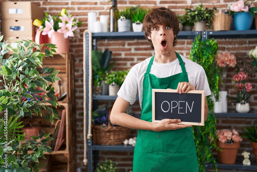 Hispanic young man working at florist holding open sign afraid and shocked with surprise and amazed expression, fear and excited face. Hispanic young man working at florist holding open sign afraid and shocked with surprise and amazed expression, fear and excited face.
