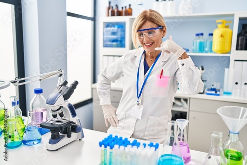Middle age blonde woman working at scientist laboratory smiling doing phone gesture with hand and fingers like talking on the telephone. communicating concepts.
