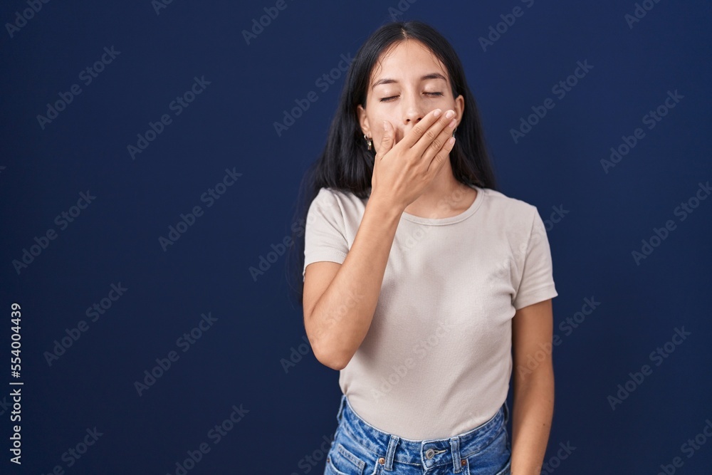 Young hispanic woman standing over blue background bored yawning tired covering mouth with hand. restless and sleepiness. Young hispanic woman standing over blue background bored yawning tired covering mouth with hand. restless and sleepiness.