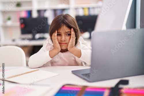 Adorable hispanic girl student using laptop with worried expression at classroom