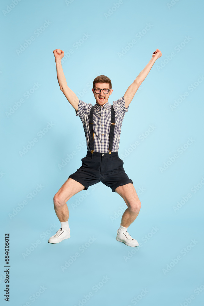 Obraz premium Portrait of young man in checkered shirt, shorts and suspenders showing happiness and excitement isolated over blue backgrounds