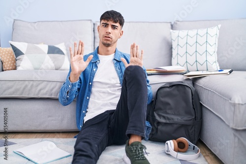 Young hispanic man sitting on the floor studying for university moving away hands palms showing refusal and denial with afraid and disgusting expression. stop and forbidden. photo