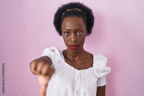 African woman with curly hair standing over pink background looking unhappy and angry showing rejection and negative with thumbs down gesture. bad expression.